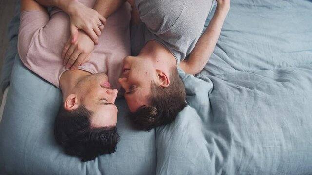 Overhead shot of loving same sex male couple lying on bed at home holding hands and kissing together - shot in slow motion