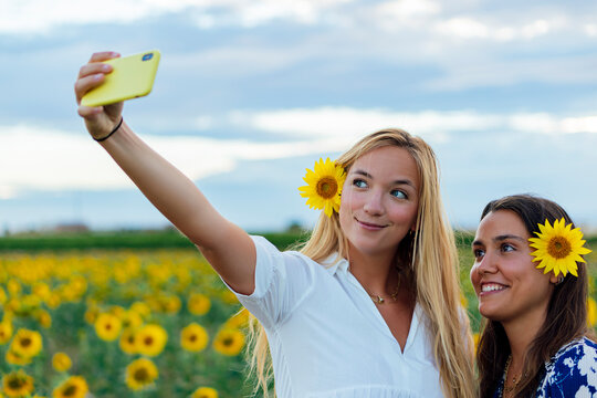 A couple of attractive young women one blonde and the other brunette posing in their designer dresses in a field of sunflowers using their smartphone