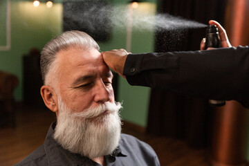 barber sprays lacquer on the hair of a man with a beard
