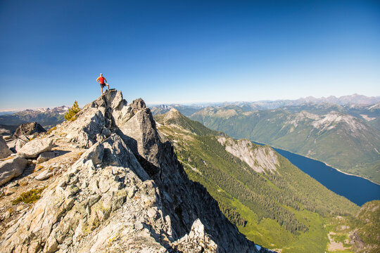 Hiker standing on mountain summit with view of lake and forest below. - Powered by Adobe