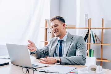 Smiling businessman having video chat on laptop near headphones and papers on blurred foreground