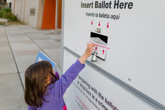 Child wearing face mask placing ballot in absentee voting drop box