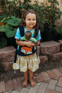 Preschool aged girl carrying babydoll in baby carrier outside in garde