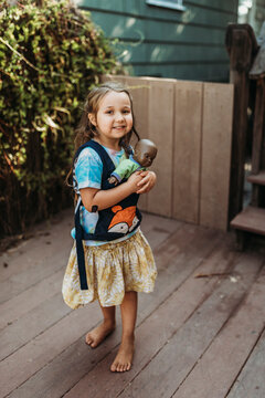 Little girl carrying babydoll in baby carrier outside in yard