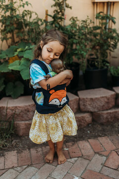 Preschool aged girl cuddling babydoll in baby carrier outside in garde