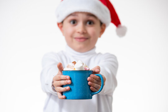 A boy in a red santa hat smiles and shows the camera a blue cup with marshmallows. Christmas concept.