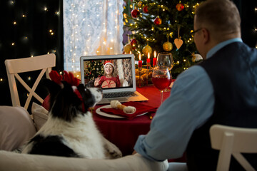 Mature man celebrating Christmas with his dog sitting at served holiday  table with laptop. People greeting their friends on video call using webcam. Christmas eve online. New normal social distancing