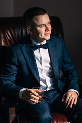 Wedding portrait of a stylish groom holding a glass of whiskey. The businessman is resting in the house and drinking alcoholic beverages. A handsome and smiling man is sitting on the couch.