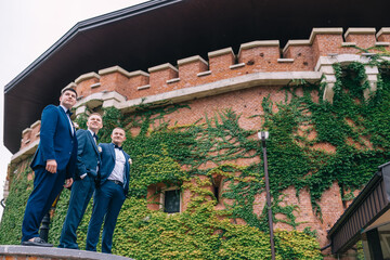 a handsome groom with his friends are standing near the castle covered with green foliage, waiting for the bride.