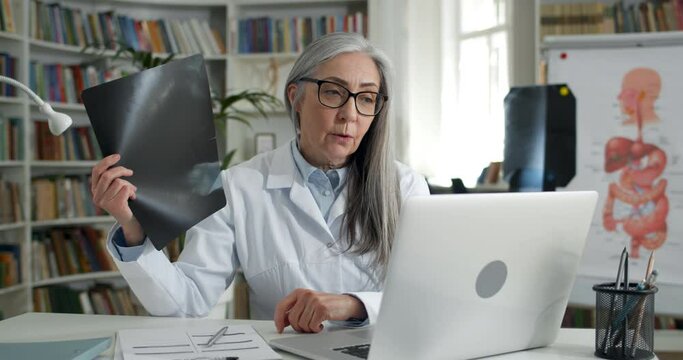 Crop view of female doctor showing x ray photo while having online medical consultation in office. Mature woman in glasses and white rob using laptop while communicating with patient