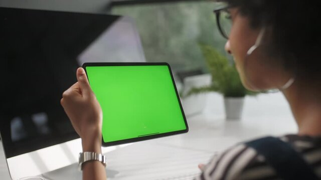 View From the Shoulder of Woman Holding and Using Hand Gestures Swipe Up on Green Mock-up Screen Digital Tablet Computer Sitting at The Desk. Browsing the Internet. Background Office.