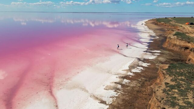 cinematic aerial 4k footage from drone flying over amazing landscape with natural pink lake, blue sky and clouds, white coast from dry salt and tourists walking along coastline
