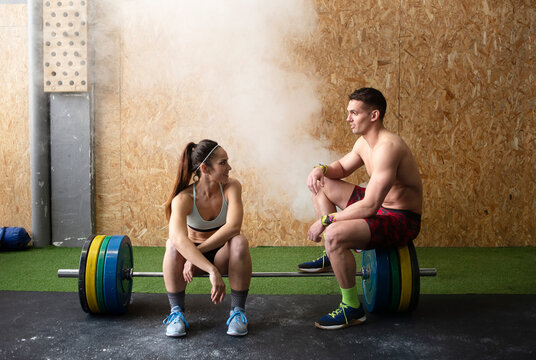 Strong shirtless man and woman in sportswear smiling and having nice conversation while sitting on barbell in gym