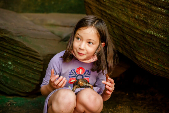 A smiling little girl crouches on ground with a painted turtle in lap