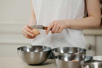 A photo of the hands of a young woman who is breaking the eggshell of the organic farm egg with the knife above the stainless steel soup bowl in the kitchen