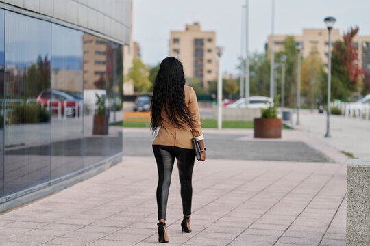 Back view of a black woman wearing a brown suit holding a laptop and a container of coffee walking next to a building. business concept