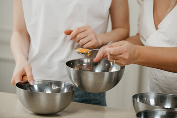 A close photo of the hands of two women, one of them is moving the yolk in a spoon to another stainless steel soup bowl in the kitchen.