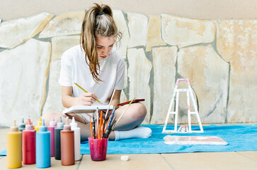 white girl painting a picture sitting on a terrace with all painting elements around as brushes, paintings, water, easel..