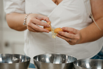 A close photo of the hands of a young woman who is separating the yolk from the egg white above the stainless steel soup bowl in a kitchen.