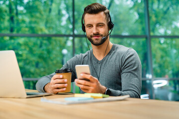 Smiling good-looking 30s man working with headset and phone at office desk holding cup of coffee