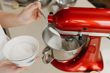 A close photo of the hands of a young woman who is spooning sugar to stainless steel soup bowl with the egg whites while the red stand mixer is whipping it.