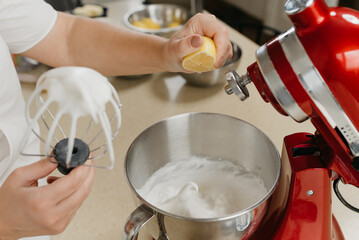 A photo of the hand of a woman who is crushing lemon juice to the mixed meringue in a stainless steel soup bowl in a kitchen.