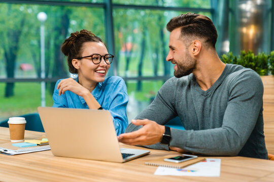 Two Young Caucasian Colleagues Talking At Office Desk Using Laptop