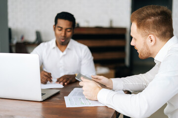 Confident happy young multiethnic businessmen in formalwear sitting together at office desk and having business team meeting in modern conference room,