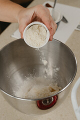 A close photo of the hands of a young woman who is tossing an almond flour from the cup into the stainless steel bowl of the mixer.