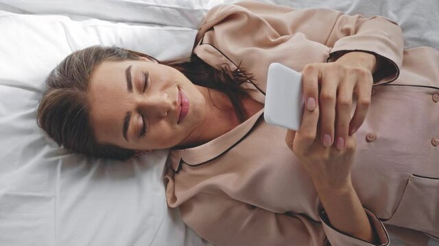 cheerful woman chatting on smartphone while lying in bed