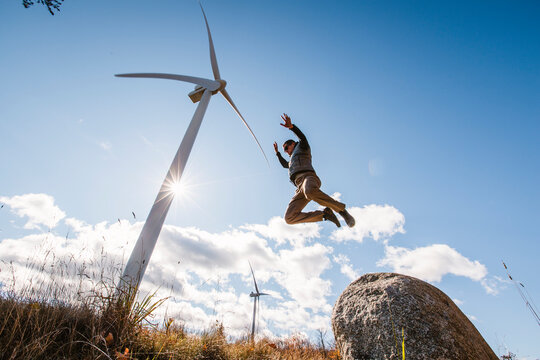 man jumps from boulder with wind turbine towering in background