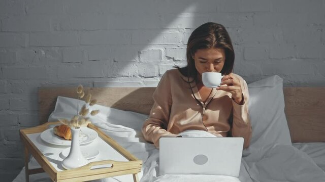 young freelancer drinking coffee and typing on laptop in bed
