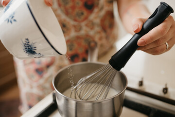 A close photo of a hand of a young woman who is mixing by the corolla cream, crushed zest, lemon juice and adding sugar into the saucepan on the gas stove