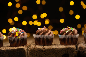 Chocolate cupcakes with sprinkling on wooden table on black background with yellow lights. Sweet dessert. Bakery concept. Elegant food. Sweets for coffee or tea. Space for text.