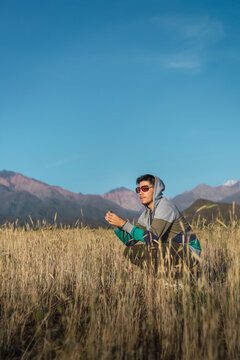 young man with sunglasses sitting on in field looking at mountains wit