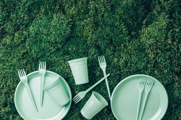 Environmental pollution. Disposable plastic tableware - plastic plates, forks, spoons, knives. Pastel plastic tableware on green grass, moss background. Top view. Copy space. Ban single use plastic