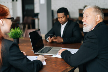 Skilled mature adult male businessman having meeting with young diverse multi-ethnic business colleagues in modern office. Multiethnic business team of businessmen people discussing new project.