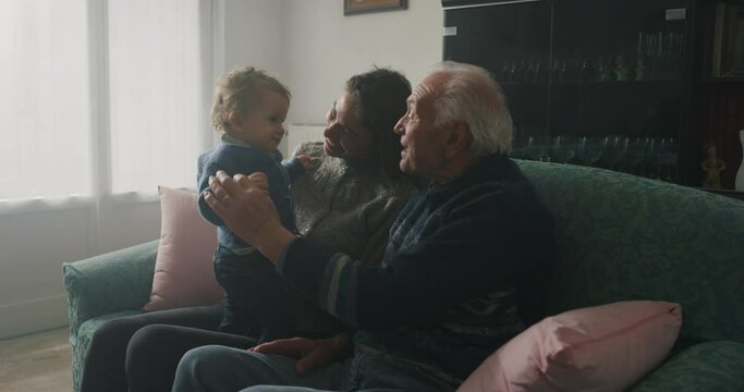 Cinematic shot of happy family: grandfather, daughter and grandson baby are having fun to pass time together while sitting on sofa at home. Concept of life, grandparents, generation, childhood, love