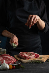 Close-up view of chef in black uniform pours rosemary to raw steak on wooden chopped board. Backstage of preparing grilled pork meat at restaurant kitchen on dark blue background. Frozen motion.