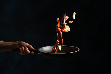 Close-up view of the chef hand toss up crab legs with garlic above the frying pan on black background. Backstage of cooking delicious meal for dinner. Seafood concept. Frozen motion. Asian cuisine.
