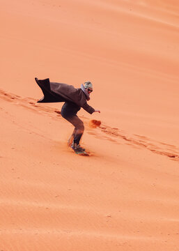 Young Man Sand Dune Surfing Wearing Bisht - Traditional Bedouin Coat. Sandsurfing Is One Of The Attractions In Wadi Rum Desert