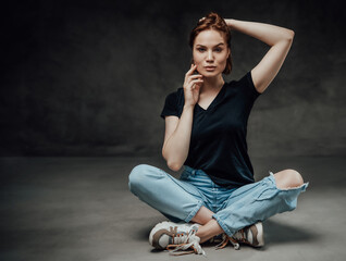 Fototapeta na wymiar Glamour and gentle female model in black shirt and ripped jeans poses sitting on floor and holding her hairs in dark background.