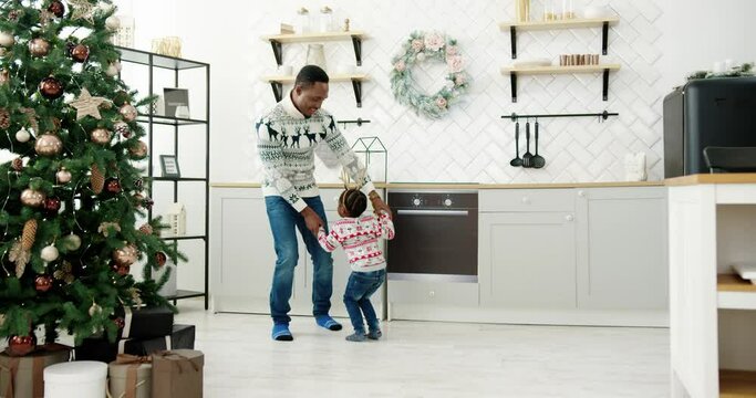 Happy African American dad and little kid listen music and dancing in modern cozy house kitchen decorated with xmas tree twinkle lights on Christmas Eve. Happy family enjoy free time, holidays concept