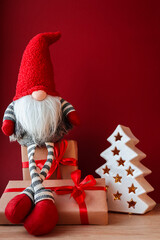 A dwarf sits on gift boxes on a red background near a Christmas tree with a garland