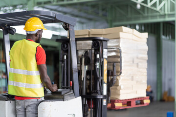 African American male warehouse worker in safety vest and helmet driving and operating on forklift...