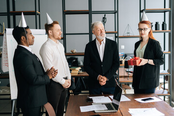 Happy business team of multiethnic businessmen people giving present to smiling mature adult male leader. Creative multiethnic business team wearing festive holiday hats having fun together.