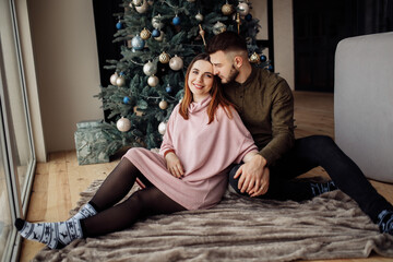 Obraz premium Adorable couple sit near the Christmas tree at home, smiling, charming husband hugging gorgeous wife, enjoy winter holidays together, New Year celebration concept