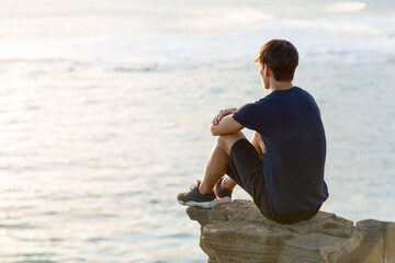 Thoughtful young man sitting alone on top of a cliff above the ocean view at sunset. - Powered by Adobe