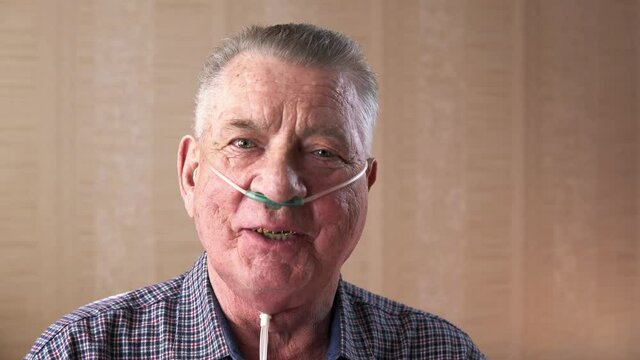 Portrait of talking senior man wearing nasal oxygen tube who has chronic obstructive pulmonary disease. An elderly person with respiratory catheter is sitting in a room. Concept of medicine and health