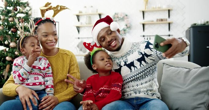 Portrait of happy African American family taking pictures on smartphone and smiling in cozy christmassy decorated home. Dad in santa hat taking selfie photo on cellphone with kids and wife